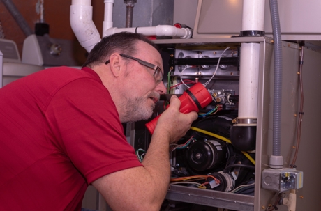 A technician wearing a red shirt inspects a furnace using a red flashlight