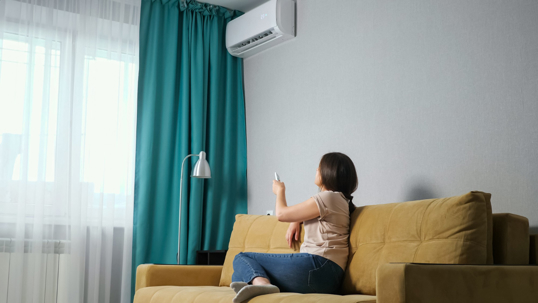 A woman sits on a couch and uses the remote to turn on an air conditioner.
