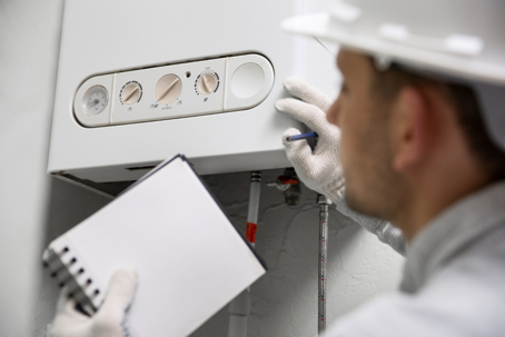 A technician in a hardhat inspects a white boiler, checking its settings, while holding a notepad and pencil