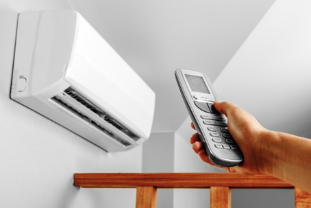 An air conditioner is mounted to the wall, and a hand holds the remote.