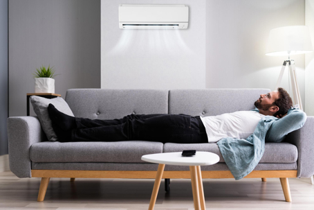 A man lounges on a couch, enjoying the cool air from a wall-mounted air conditioner.