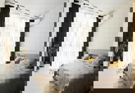 A well-lit bedroom features soft colors, sheer white curtains with dark drapes, and a bed covered with a gray quilt and yellow accent pillows.