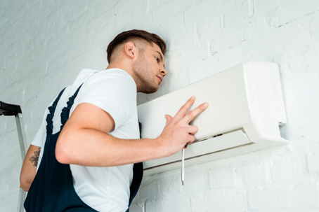 Technician fixing an air conditioner. A handyman in a white shirt and blue coveralls repairs an air conditioning unit on a white brick wall with a screwdriver.