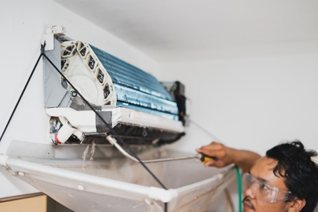 A person cleans an air conditioner with a pressurized hose while wearing safety glasses.