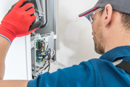 A technician wearing safety glasses and work gloves is repairing the electrical components inside a heating appliance.