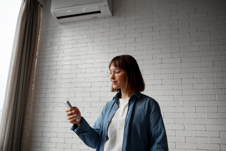 A woman points a remote at an air conditioner mounted high on a white brick wall.