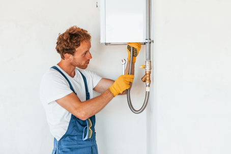 Plumber in overalls, wearing gloves, uses a wrench on pipes connected to a wall-mounted water heater.