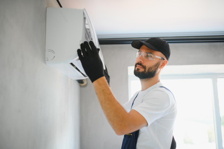 A technician wearing safety glasses and gloves is seen servicing an air conditioner unit mounted on a wall.